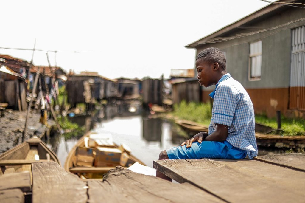 A young boy in a village sits alone thoughtfully by a canal, reflecting on his surroundings.